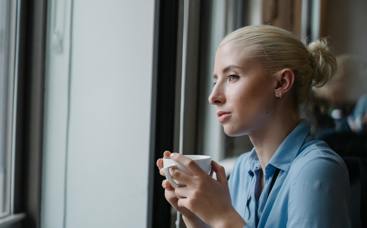 a young woman sitting in a cafe with a coffee in hand, looking outside the window. 